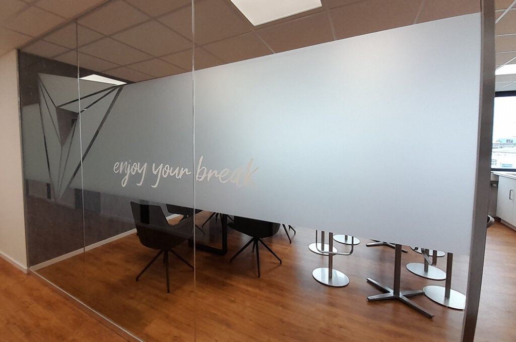 Besprechungsraum mit Glastrennwand, Aufschrift "Enjoy your break", Holzboden, Sonnenlicht, leere Stühle.