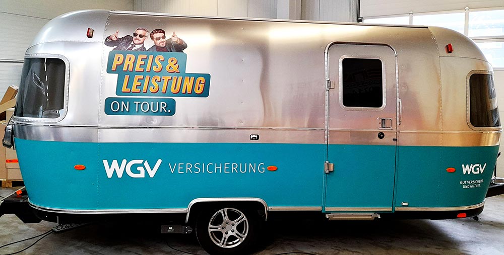 Silberner und tuerkiser WGV Wohnanhaenger mit Werbegrafik und Gesichtern, freundlich schauend